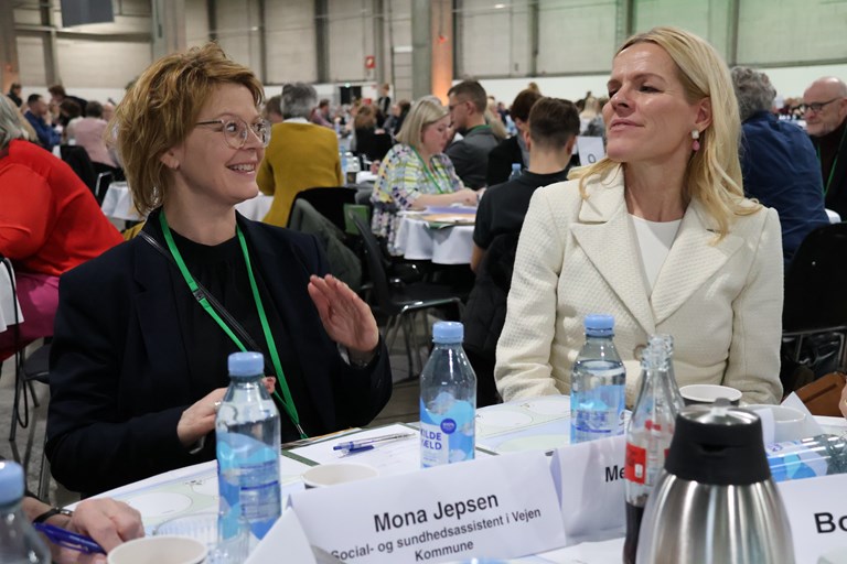 Billedet viser ældreminister Mette Kierkgaard (til højre) og social- og sundhedsassistent, Mona Jepsen (til venstre) talende og siddende ved et bord.